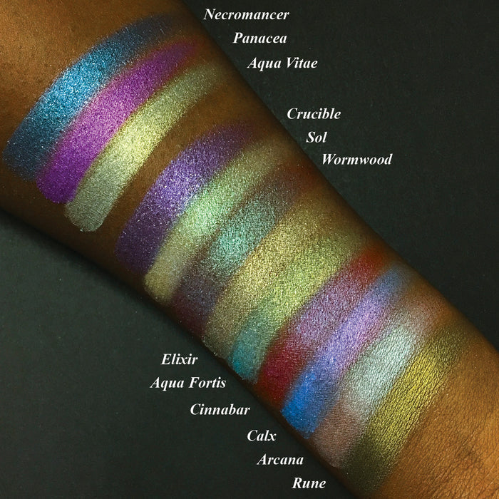 Top angled arm swatches on deep skin tone Witchcraft vs. Alchemy Collection including Crucible Duochrome Eyeshadow next to Necromancer, Panacea, Aqua Vitae, Sol, Wormwood, Elixir, Cinnabar, Calx, Arcana, Rune, Aqua Fortis