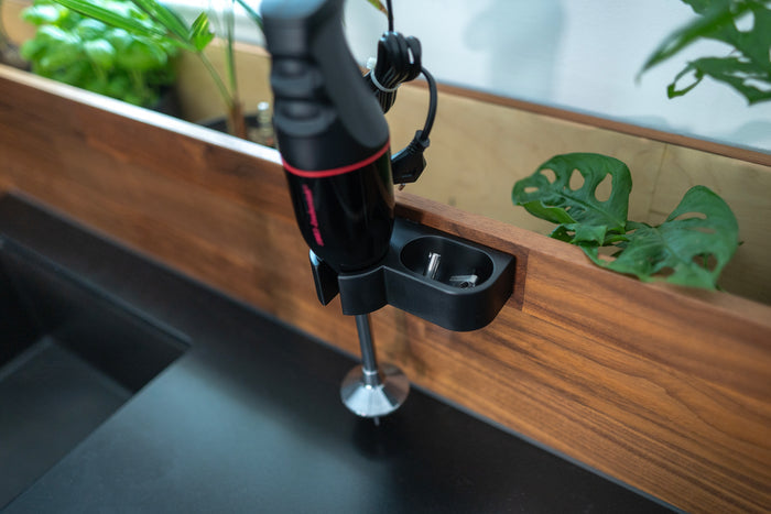 Der Stabmixer Unold ESGE-Zauberstab® M 100 Design Schwarz ist auf einem Holzgestell über einer dunklen Küchenarbeitsplatte montiert, im Hintergrund sind grüne Topfpflanzen zu sehen.