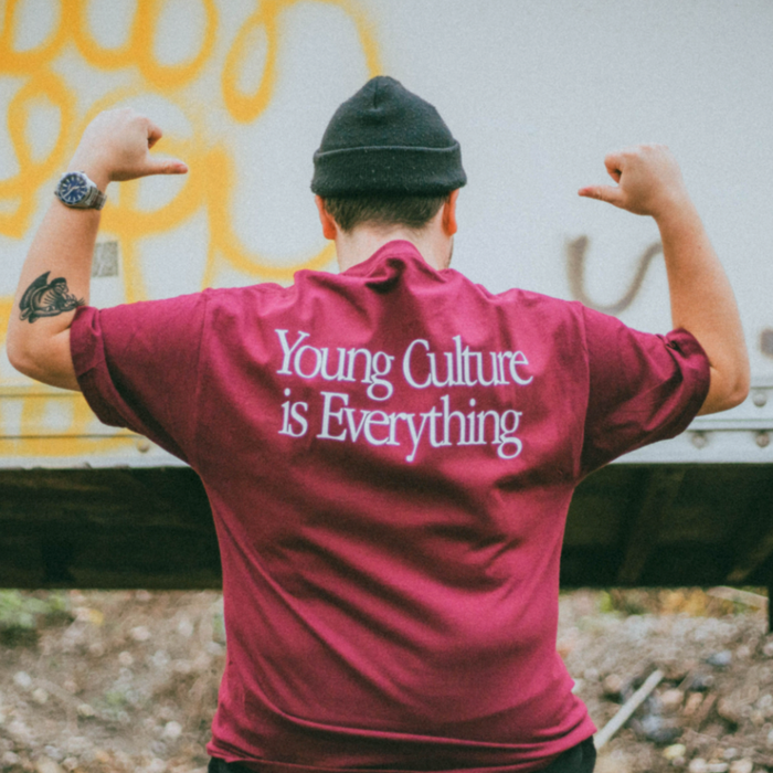 A person wearing a maroon Young Culture t-shirt with “Young Culture is Everything” on the back flexes for the camera, paired with a black beanie and showing off a left arm tattoo—an ideal look for fall.