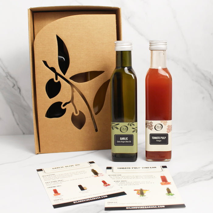 Two bottles of gourmet food items, one labeled as Garlic Olive Oil and the other as Tomato Pulp Vinegar, are presented alongside their packaging box with a leaf design. There are also informational cards in front of the bottles.