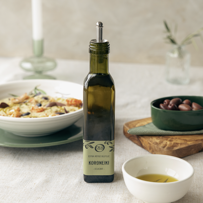 Koroneiki extra virgin olive oil on a white table next to a bow of olives and a small bowl of olive oil