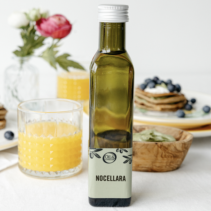 Nocellara Extra virgin olive oil bottle on a white table next to orange juice in a glass and other breakfast foods