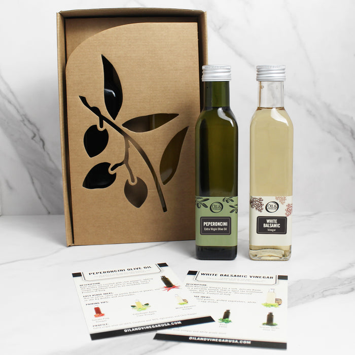 A bottle olive oil and white balsamic vinegar placed outside a cardboard box with a leaf design. The vinegar bottle is labeled with 'White Balsamic' and the olive oil bottle with 'Peperoncini Extra Virgin'.