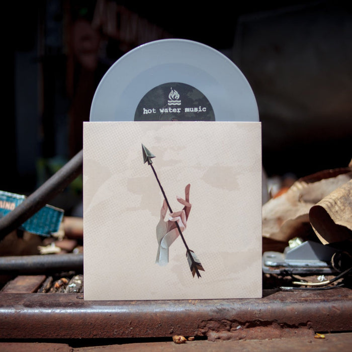 A Hot Water Music vinyl record in a translucent sleeve stands behind an album cover with an abstract hand holding a spear, displayed outdoors on rustic metal—part of the Remnants/Fences release.