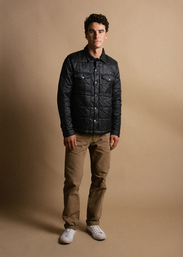 Man in a quilted black jacket, khaki pants, and white sneakers stands against a beige background, looking confidently at the camera.