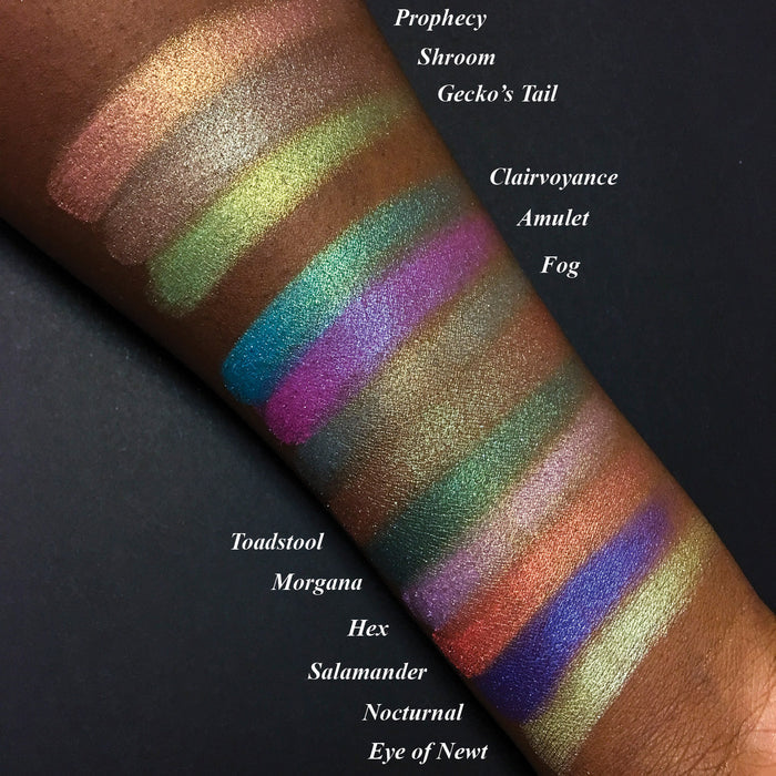 Top angled arm swatches on deep skin tone of Witchcraft vs. Alchemy Collection including Salamander Duochrome Eyeshadow next to Prophecy, Shroom, Gecko's Tail, Clairvoyance, Amulet, Toadstool, Morgana, Hex, Nocturnal, Eye of Newt, Fog