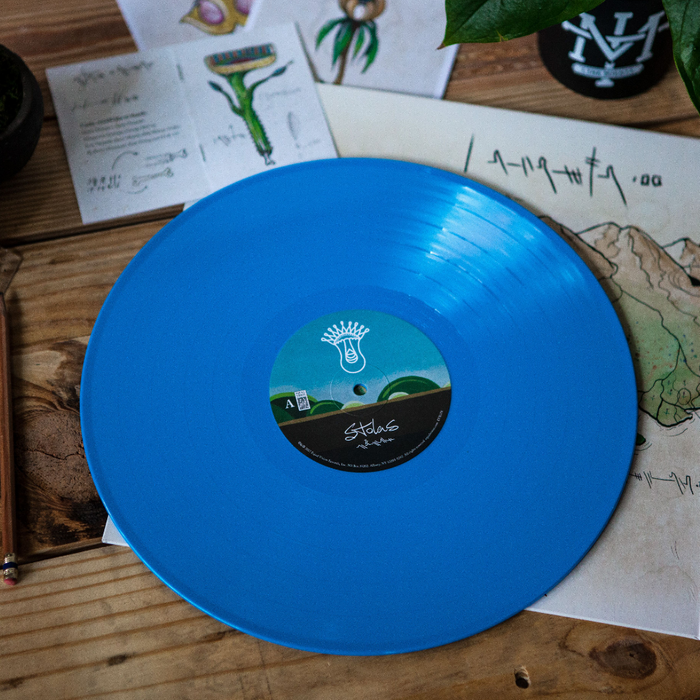A vibrant opaque blue Stolas Self-Titled vinyl record rests on a wooden table, surrounded by illustrated papers, a pen, a black mug with a white design, and a green plant in the corner.