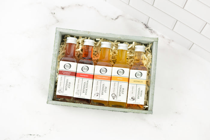 A gift set of five 100ml vinegar bottles in a wood tray with shredded paper filler, displayed on a white marble surface with a white subway tile background.