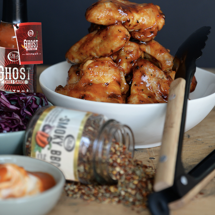 Bowl of chicken wings with a ar of smoky BBQ seasoning lying on its side in front of the bowl next to black tongs. Ghost chili sauce is in the background
