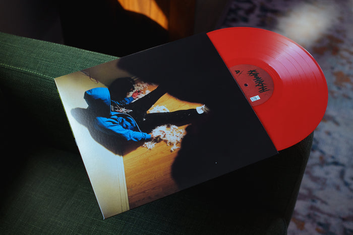 A red vinyl peeks out of a jacket with a picture of a man with a blue hoodie and dreads slumped over against a wall with a dark figure over him. The vinyl and jacket rest against a green couch.