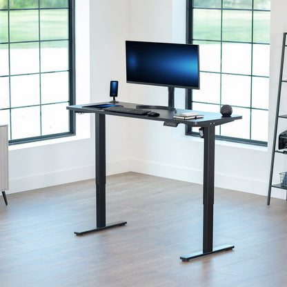 Black adjustable desk with computer setup in a bright room with large windows.