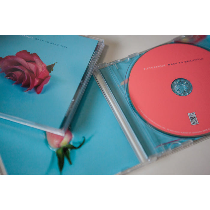 There is a square CD with a blue background and a red rose in the middle that is half submerged in the blue background located in the left corner of the image. The CD case is open in the image to show that the color of the cd is bright pink.