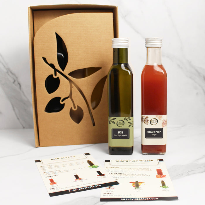 Two bottles of gourmet food items; one labeled as Basil Olive Oil and the other as Tomato Pulp Vinegar, presented next to a cardboard box with a silhouette of olive branches.