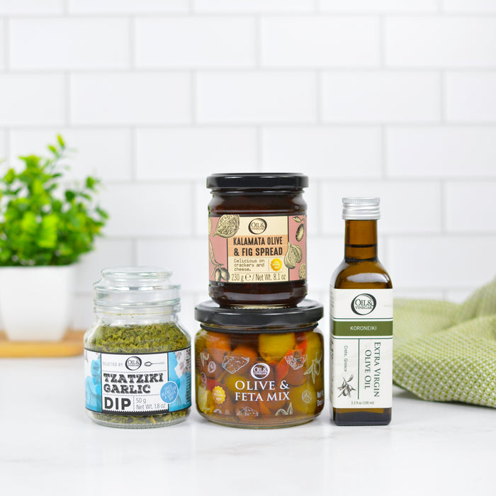 Assorted products including a jar of tzatziki garlic dip, a bottle of olive oil, Kalamata olive and fig spread, and a jar of olive and feta mix on a white surface with a tiled wall background.