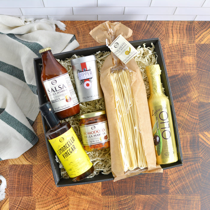 Gourmet gift box with various items including a bottle of tomato sauce, a dipper, a jar of pomodoro, a bottle of lemoncello vinegar, and a package of pasta on a wooden surface.