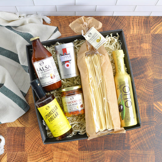 Gourmet gift box with various items including a bottle of tomato sauce, a dipper, a jar of pomodoro, a bottle of lemoncello vinegar, and a package of pasta on a wooden surface.