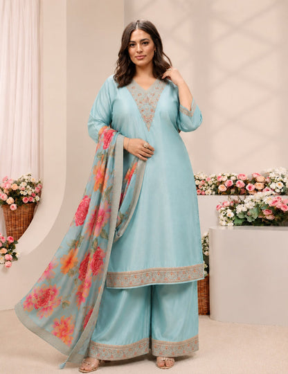 A woman modeling a light blue three-piece ethnic suit with matching floral dupatta in a styled indoor studio setting with flowers.