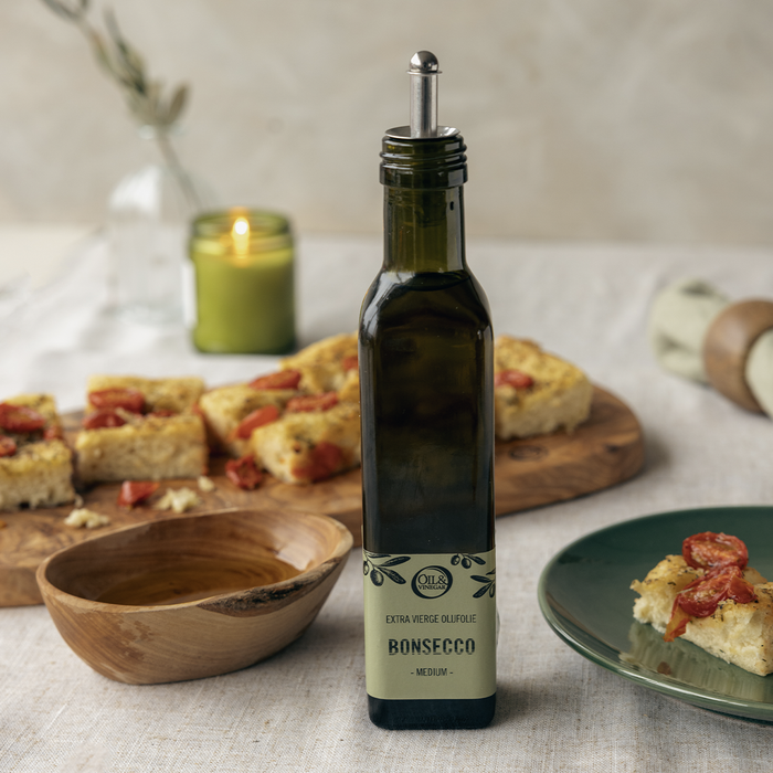 Bottle on Bonsecco extra virgin olive oil on a linen table cloth next to a tomato focaccia bread and an olive wood bow with olive oil