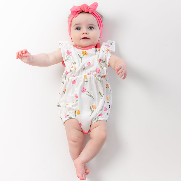 Young infant laying on a white surface wearing the Bubble Romper in Tulip paired with a pink bow