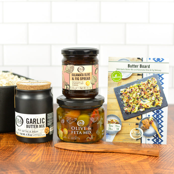 A collection of gourmet food items for making butter boards, including garlic butter mix, olive & feta mix, kalamata olive & fig spread, and an olive wood spreader, presented on a wooden surface with a box in the background.