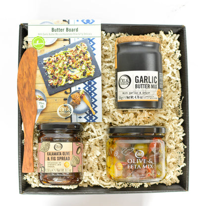 A collection of gourmet food items for making butter boards, including garlic butter mix, olive & feta mix, kalamata olive & fig spread, and an olive wood spreader, presented in a black box with crinkle paper