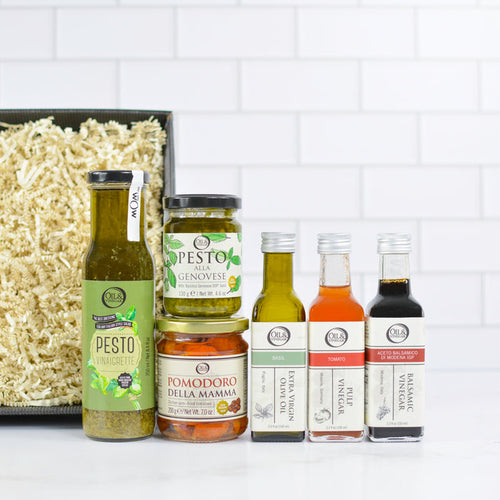 Collection of pesto, pesto dressing, pomodoro della mamma, two vinegar bottles, and one olive oil bottle on a kitchen counter with a white tiled wall background.