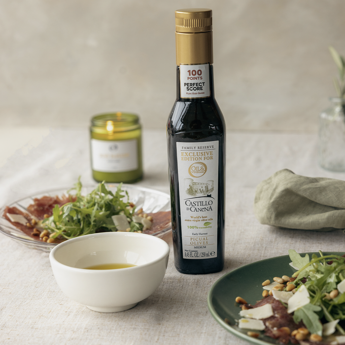 Castill De Canena extra virgin olive oil on a white table with a bowl of olive oil, a salad, a plate of carpaccio, and a candle