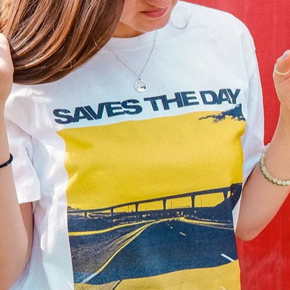 White short sleeve shirt with a yellow tinted image of a highway. There is an overpass and a cloud in the top corner. Above the image is black text that says SAVES THE DAY. An individual is modeling the shirt in front of an orange and red designed wall.