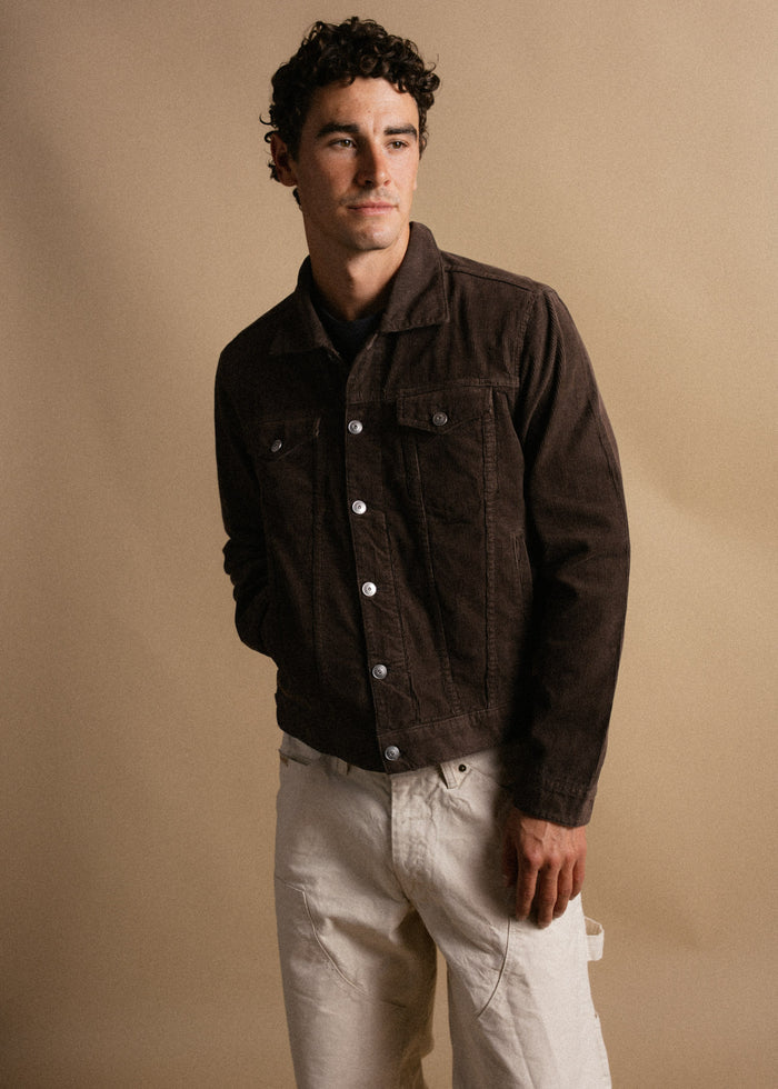 Man in a brown jacket and white pants stands against a beige background, looking to the side with a thoughtful expression.