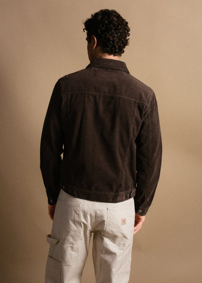 Man with curly hair in a brown jacket and beige pants stands facing away against a neutral background.