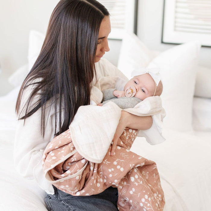 A mother holds her little girl in a Saranoni blanket. The baby is wrapped in a beautifully soft and breathable muslin quilt, designed for both comfort and style. The quilt features a warm, earthy rust tone with delicate white botanical accents, adding a timeless and elegant touch. Its airy, multi-layered muslin fabric is crafted to become even softer with each wash, making it a perfect choice for snuggles, naps, and everyday moments. The soft blanket is a luxury blanket and a Saranoni blanket.