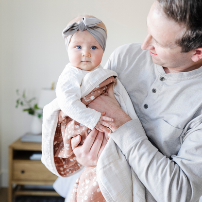 A father holds his little girl in a Saranoni blanket. The baby is wrapped in a beautifully soft and breathable muslin quilt, designed for both comfort and style. The quilt features a warm, earthy rust tone with delicate white botanical accents, adding a timeless and elegant touch. Its airy, multi-layered muslin fabric is crafted to become even softer with each wash, making it a perfect choice for snuggles, naps, and everyday moments. The soft blanket is a luxury blanket and a Saranoni blanket.
