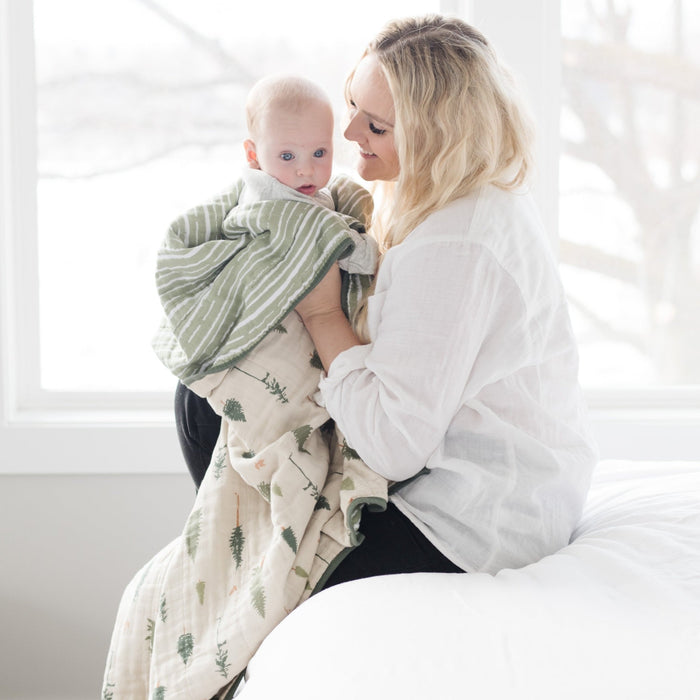 A mother holds her baby boy who is wrapped in a cotton muslin quilt that has a pine tree and green striped pattern on it. This soft blanket is a luxury blanket and a Saranoni blanket. The adorable pattern makes it a great addition to a baby's nursery or toddler's bedroom.