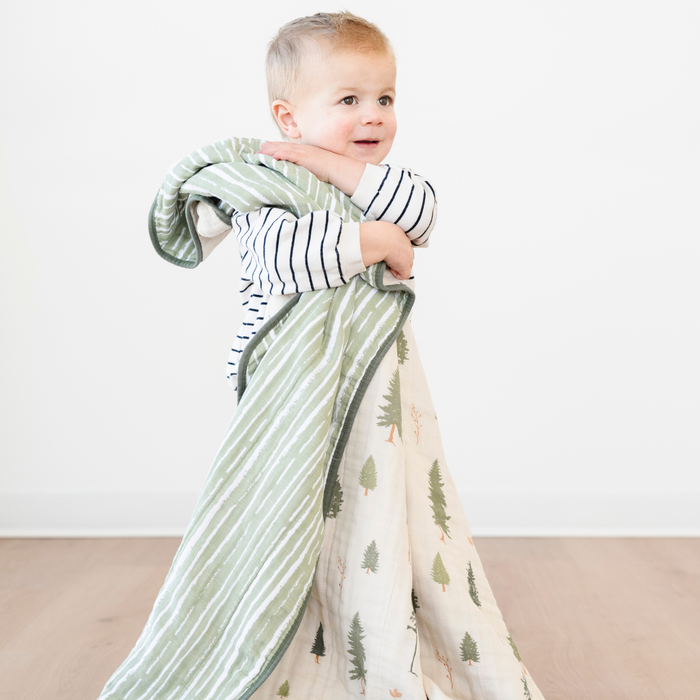 A little boy is hugging a cotton muslin quilt that has a pine tree and green striped pattern on it. This Saranoni blanket is a great toddler blanket or baby blanket. Use as an addition to a baby's nursery or toddler's bedroom.