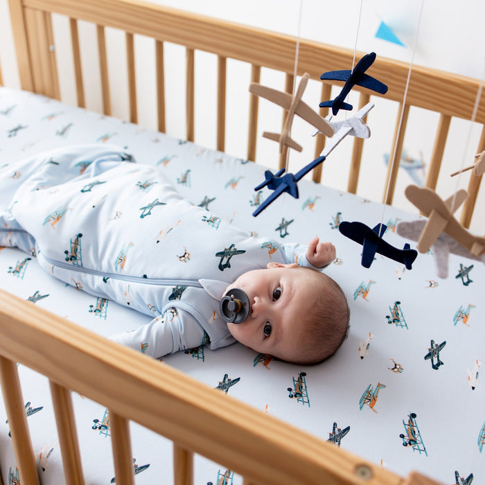 Infant laying in a crib on the Crib Sheet in Take Off wearing a matching sleep bag looking at a plane mobile hung above the crib