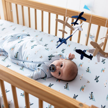 Infant laying in a crib on the Crib Sheet in Take Off wearing a matching sleep bag looking at a plane mobile hung above the crib