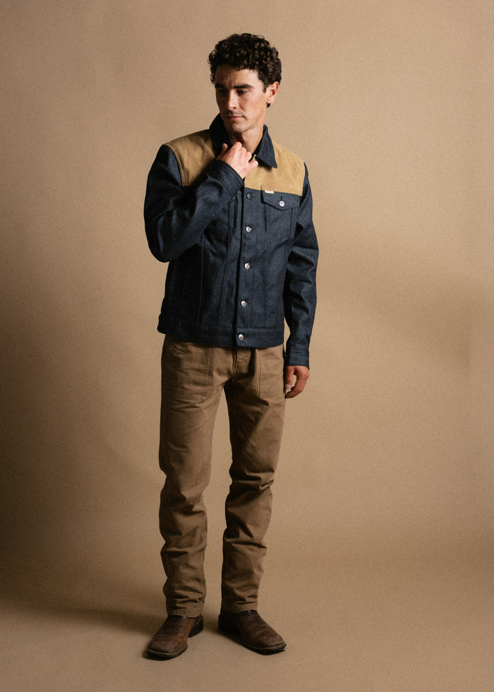 Man in a denim jacket and brown pants stands on a beige background, looking to the side with one hand on his collar.