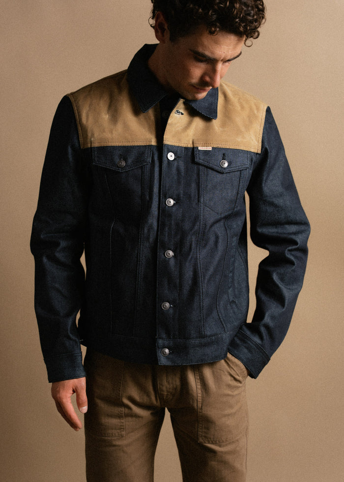 Man in a stylish denim jacket with tan accents and brown pants, looking down against a neutral background.
