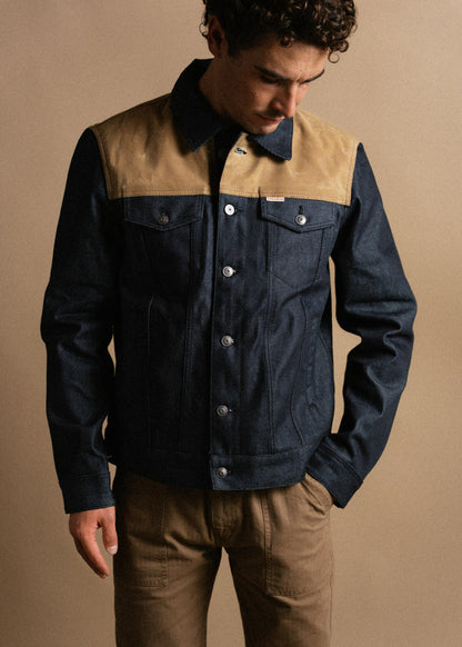 Man in a stylish denim jacket with tan accents and brown pants, looking down against a neutral background.