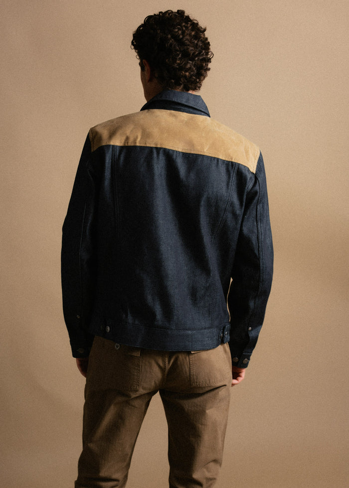 Man in denim jacket with tan shoulders, brown pants, facing away against a beige background, showcasing casual fashion style.