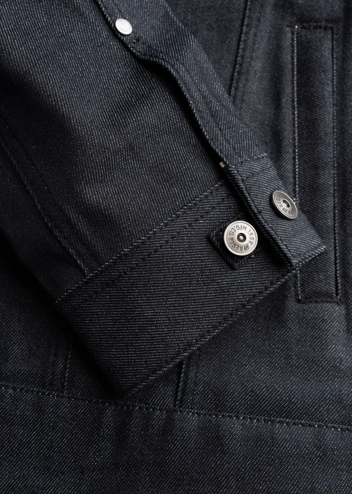 Close-up of a dark denim jacket sleeve with visible stitching and metal button details, showcasing texture and craftsmanship.