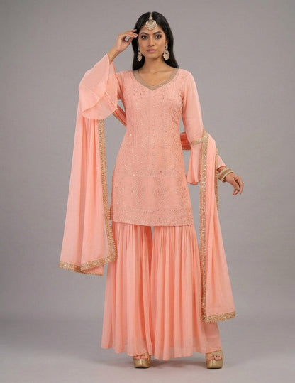 A woman modeling a peach pink traditional Indian salwar kameez suit with gold embroidery and a matching dupatta, set against a solid gray studio background.