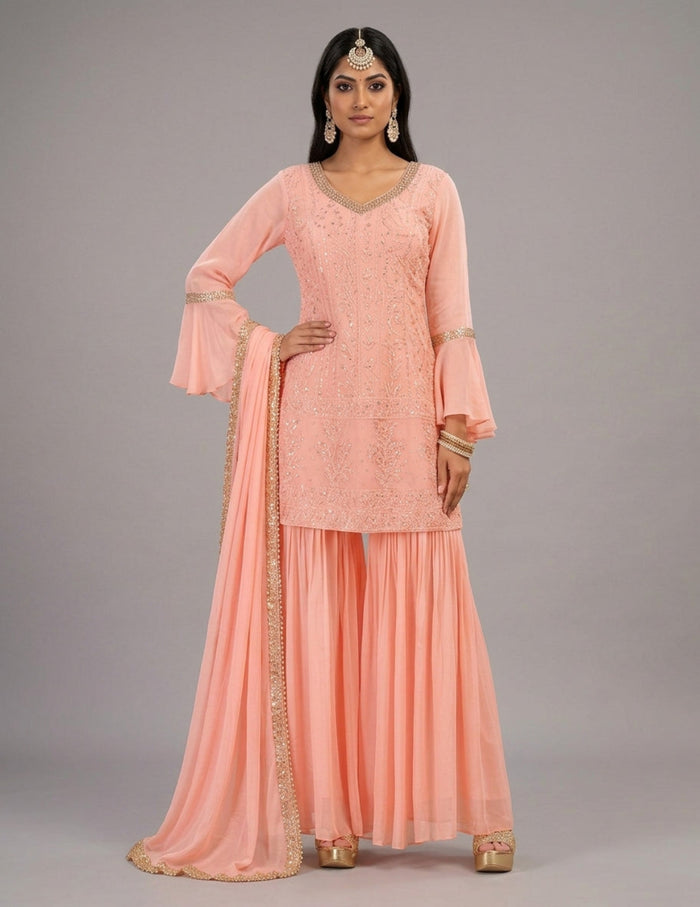A woman models a peach pink traditional Indian outfit consisting of a heavily embroidered kurta with bell sleeves, matching pleated sharara pants, and a sheer dupatta with gold zari embroidery, set against a solid gray studio background.