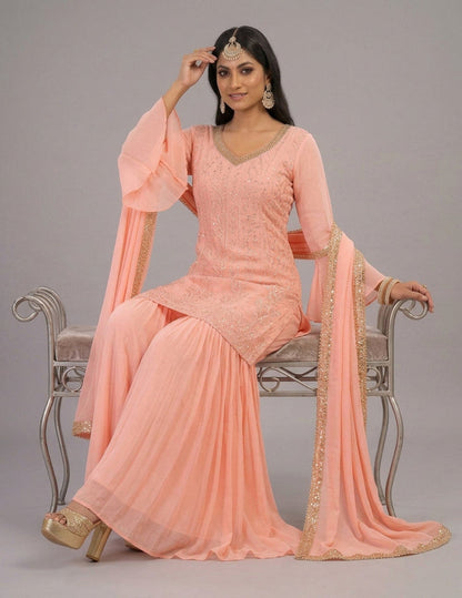 A woman models a peach pink ethnic salwar kameez suit with gold embroidery and a matching dupatta draped over a silver bench against a gray studio background.