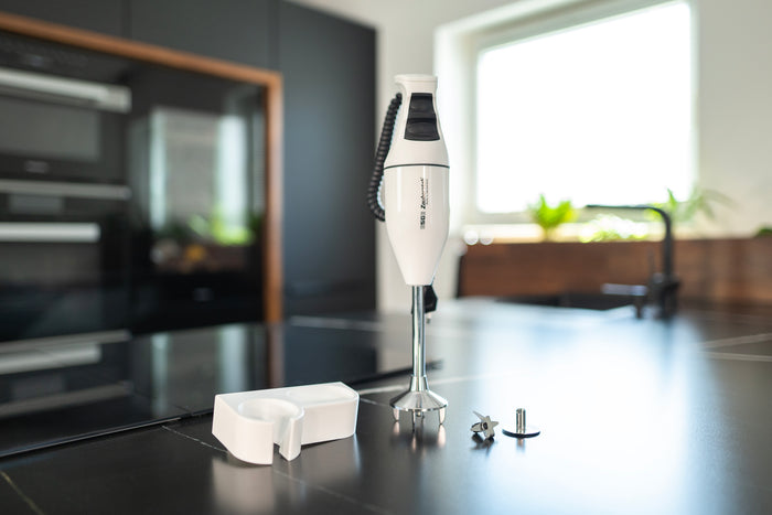 Der Unold ESGE-Zauberstab® E 120 Select Weiß, ein weißer Stabmixer mit Aufsätzen und Ständer, wird auf einer modernen schwarzen Küchentheke präsentiert, im Hintergrund ein Fenster mit Einbaugeräten.