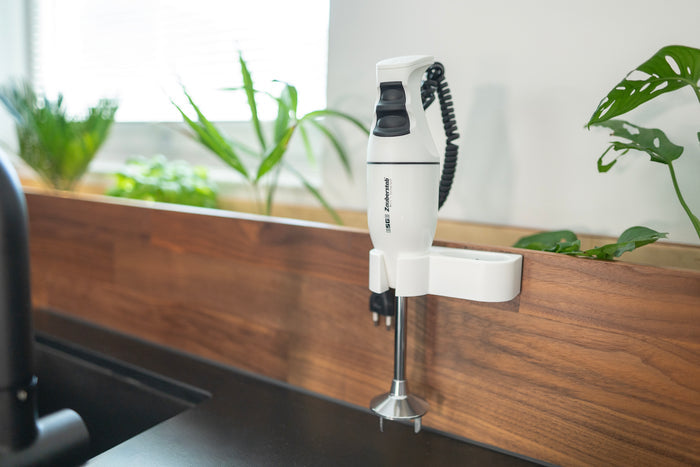 Der Stabmixer Unold ESGE-Zauberstab® E 120 Select Weiß ist auf einer Holzrückwand in einer modernen Küche montiert, im Hintergrund sind auf der Fensterbank grüne Pflanzen zu sehen.