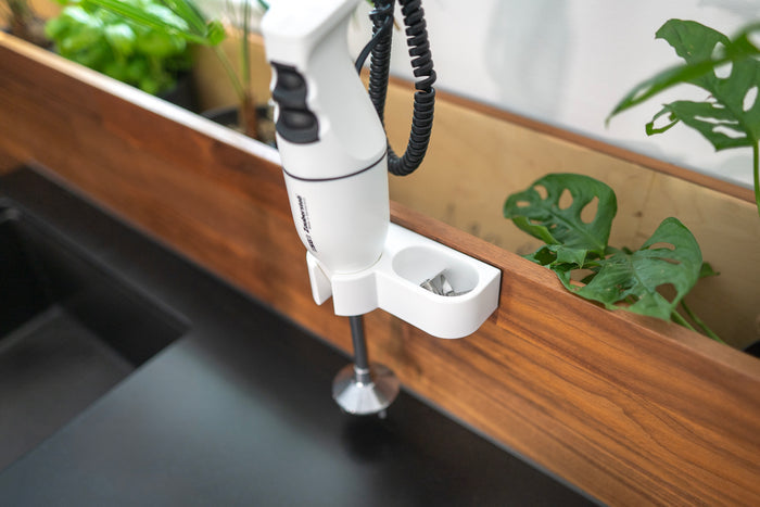 Der Stabmixer Unold ESGE-Zauberstab® E 120 Select Weiß ist auf einer Holzplatte über einer schwarzen Arbeitsplatte montiert, im Hintergrund sind grüne Topfpflanzen zu sehen.