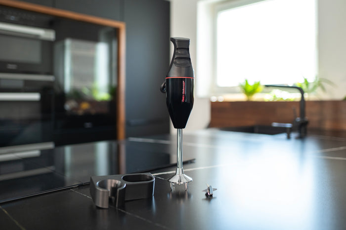 Der ESGE-Zauberstab® M 100 Design Schwarz Stabmixer von Unold inklusive Zubehör steht auf einer modernen Küchentheke. Im Hintergrund sind dunkle Schränke, eine Spüle und ein sonnendurchflutetes Fenster mit Topfpflanzen zu sehen.
