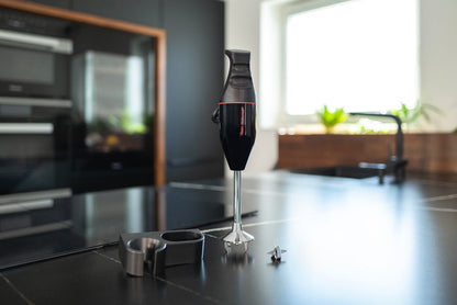 Der ESGE-Zauberstab® M 100 Design Schwarz Stabmixer von Unold inklusive Zubehör steht auf einer modernen Küchentheke. Im Hintergrund sind dunkle Schränke, eine Spüle und ein sonnendurchflutetes Fenster mit Topfpflanzen zu sehen.