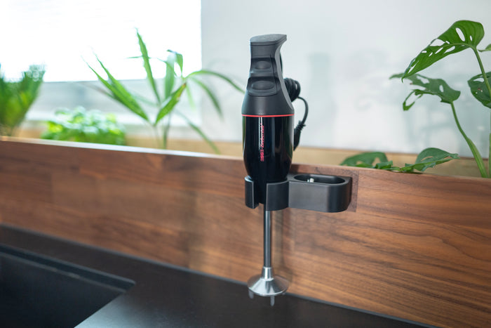 Der Stabmixer Unold ESGE-Zauberstab® M 100 Design Schwarz ist auf einer Küchenrückwand aus Holz montiert, im Hintergrund sind grüne Pflanzen und ein Fenster zu sehen.